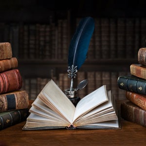 An open book with a quill and ink stand on a table surrounded by other books - Litigation Consultants